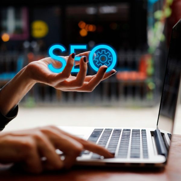 A hand holds the word SEO in front of the laptop